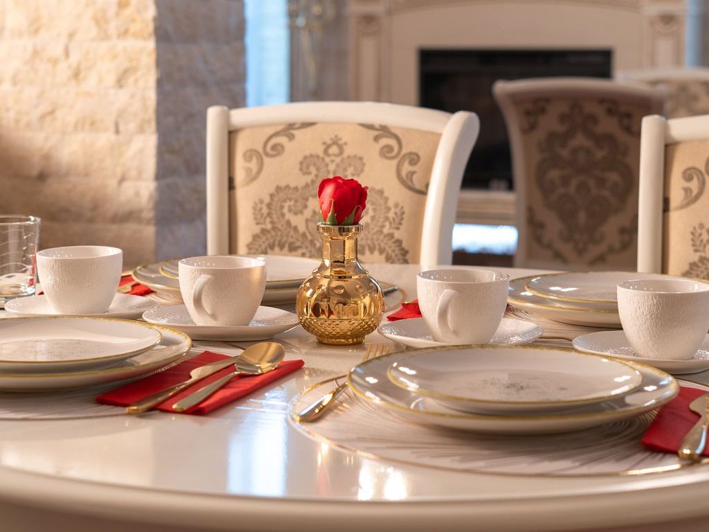 White round pedestal dining table with gold-trimmed place settings, red napkins, and a small gold vase holding a single red rose in an elegant formal dining room.