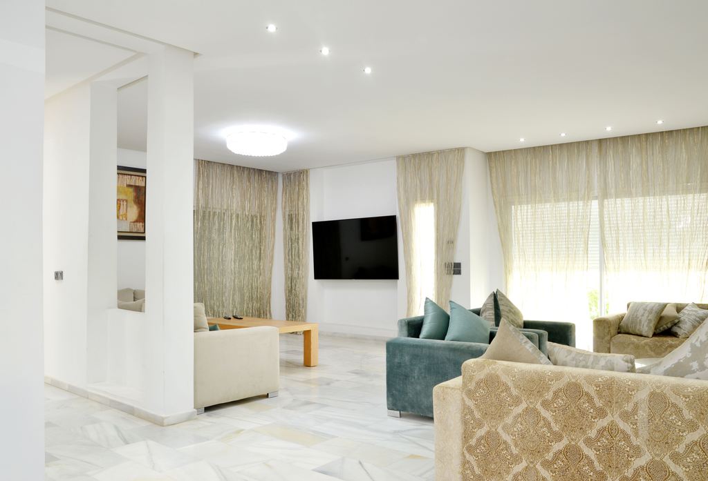 Bright open-plan home with two connected living areas, neutral sofas, a wall-mounted TV, floor-to-ceiling curtains, and white marble flooring.