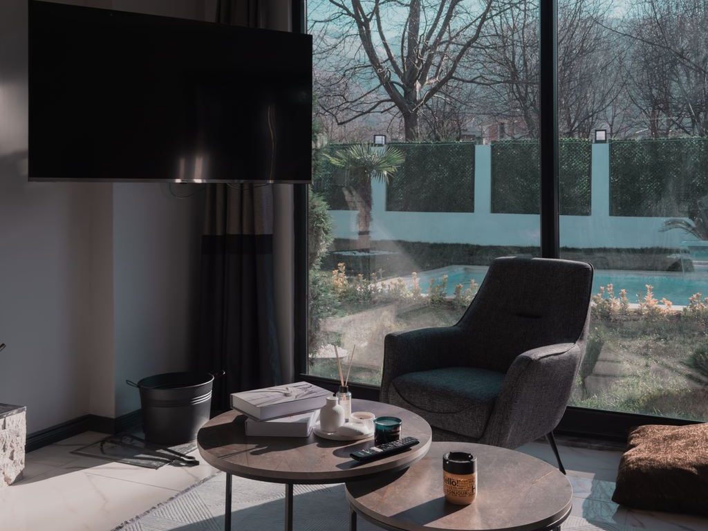 Cozy modern living room with a wall-mounted TV, dark accent chair, large windows, layered round coffee tables, and a light patterned rug.