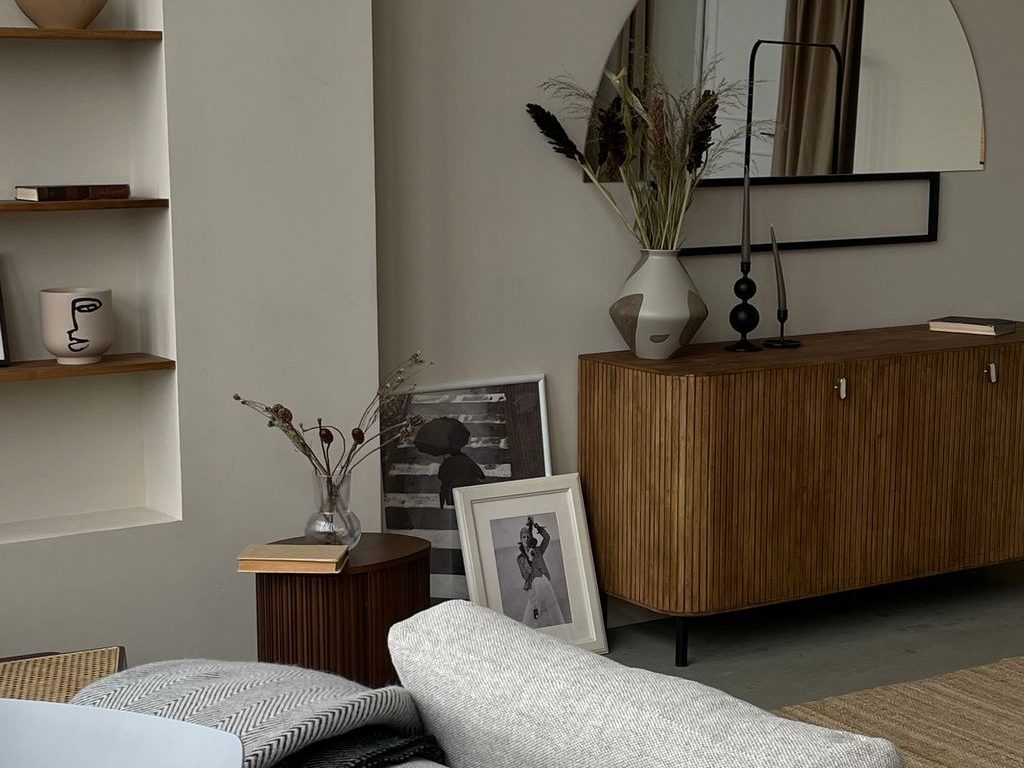 Warm living room with a gray sectional sofa, walnut fluted sideboard, curved wall mirror, built-in shelves, and neutral decorative accents.