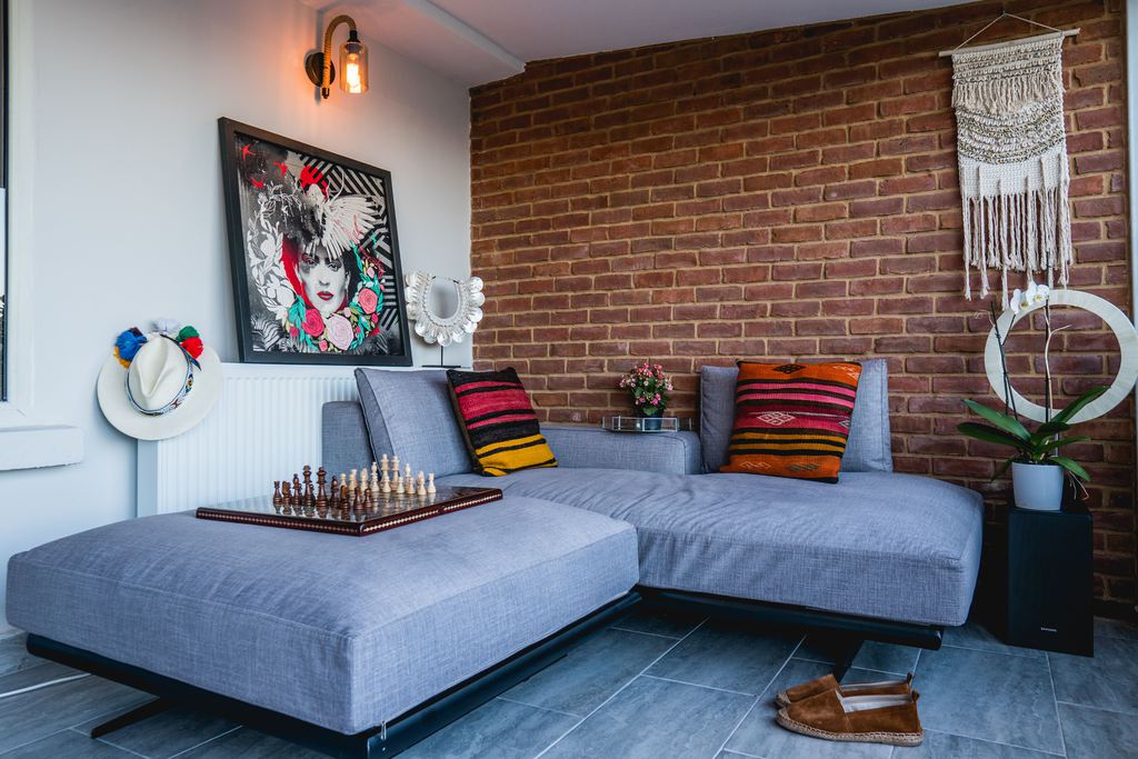 Modern eclectic living room with gray sectional sofa, brick accent wall, colorful pillows, bold portrait art, macrame wall hanging, and layered decor.