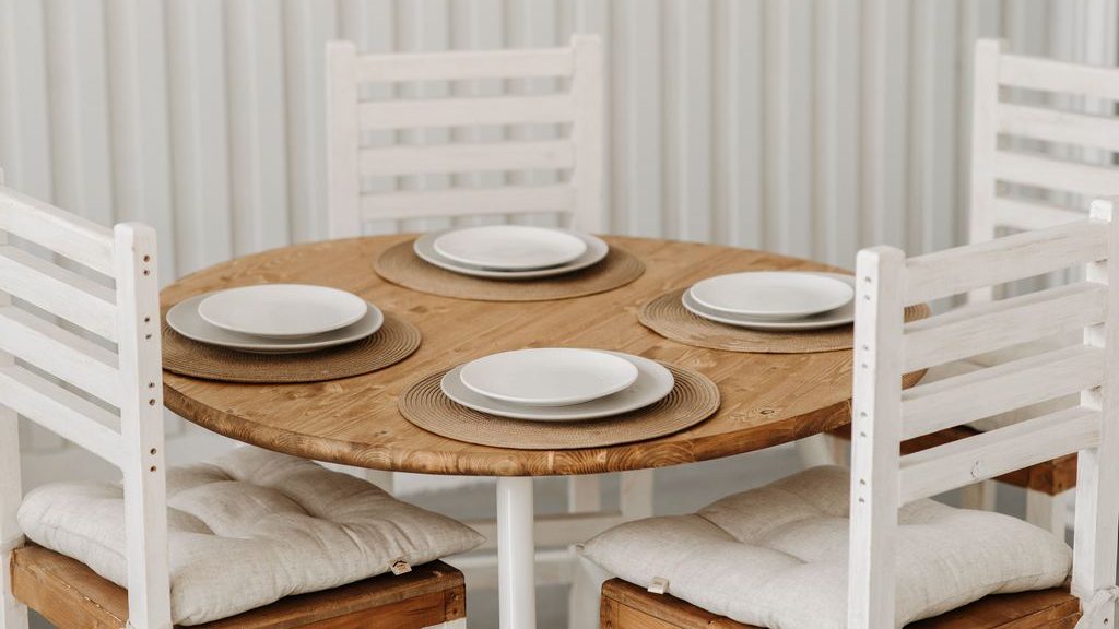 Round natural wood dining table with four white ladder-back chairs, woven placemats, and simple white place settings.
