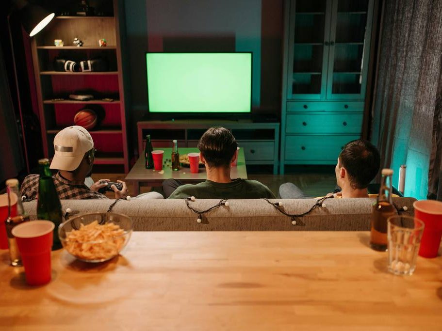 Three friends watching TV from a sofa in a small living room, with snacks and drinks arranged behind them for a casual watch party.