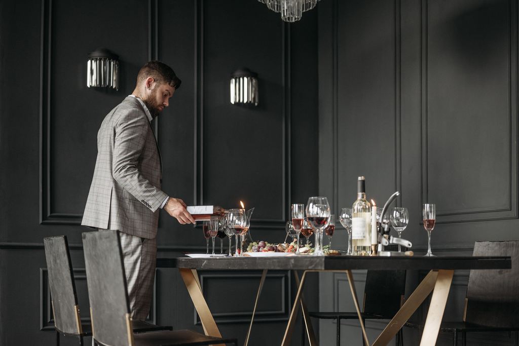 Man in a light suit pours wine at a dark dining table set with glasses, candles, grapes, and modern black wall panels.