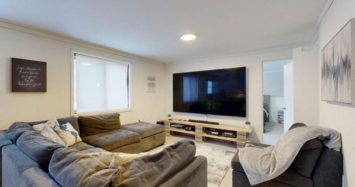 Dark gray sectional sofa and armchair facing a wall-mounted TV in a compact family room with a low media console and open walkway to an adjacent room.