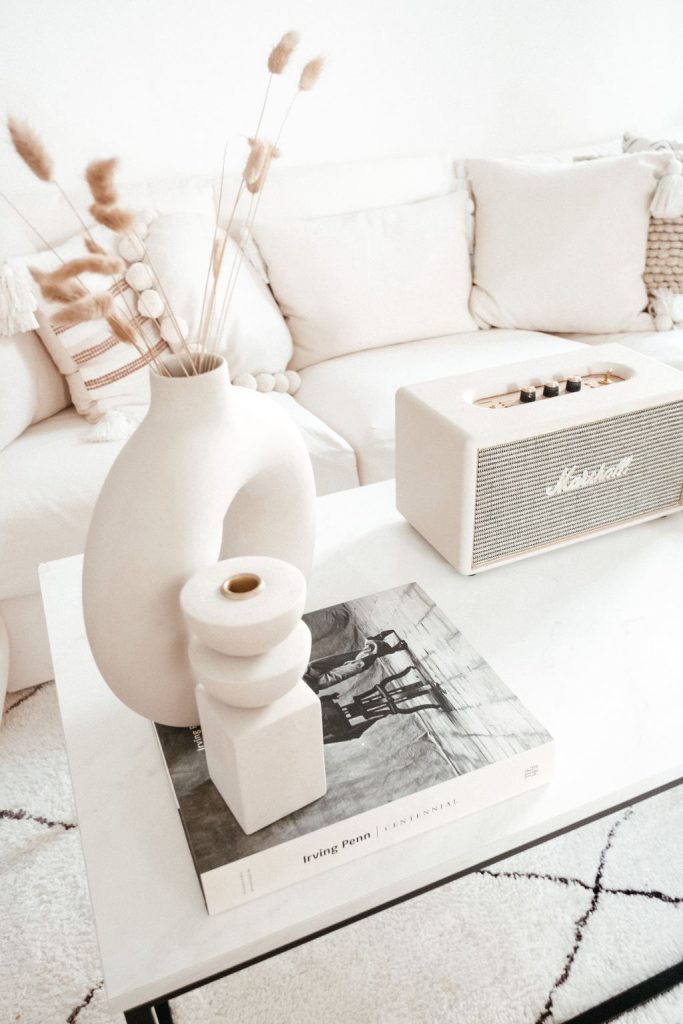 White marble coffee table styled with neutral ceramic vases, dried grasses, a design book, and a cream speaker beside a white sofa.