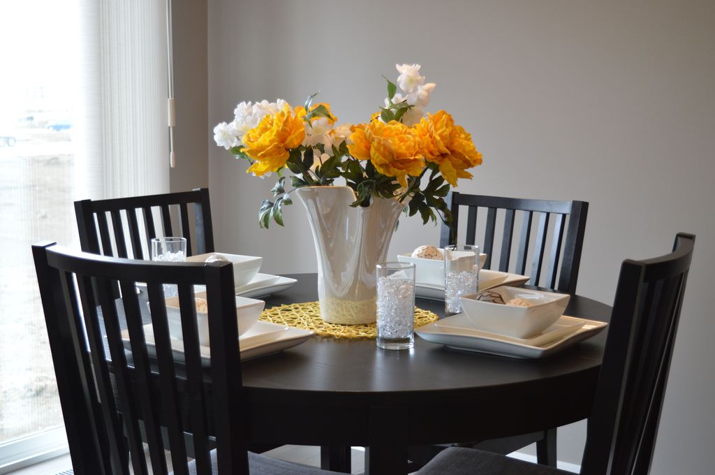 Round black dining table set with four black slat-back chairs, white place settings, and a tall white vase filled with yellow and white flowers.