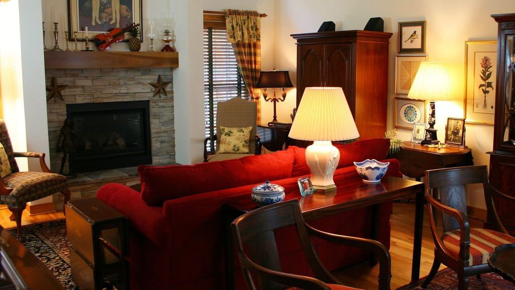 Traditional family room with a red sofa, fireplace, wood furniture, patterned armchairs, and table lamps creating warm layered lighting.