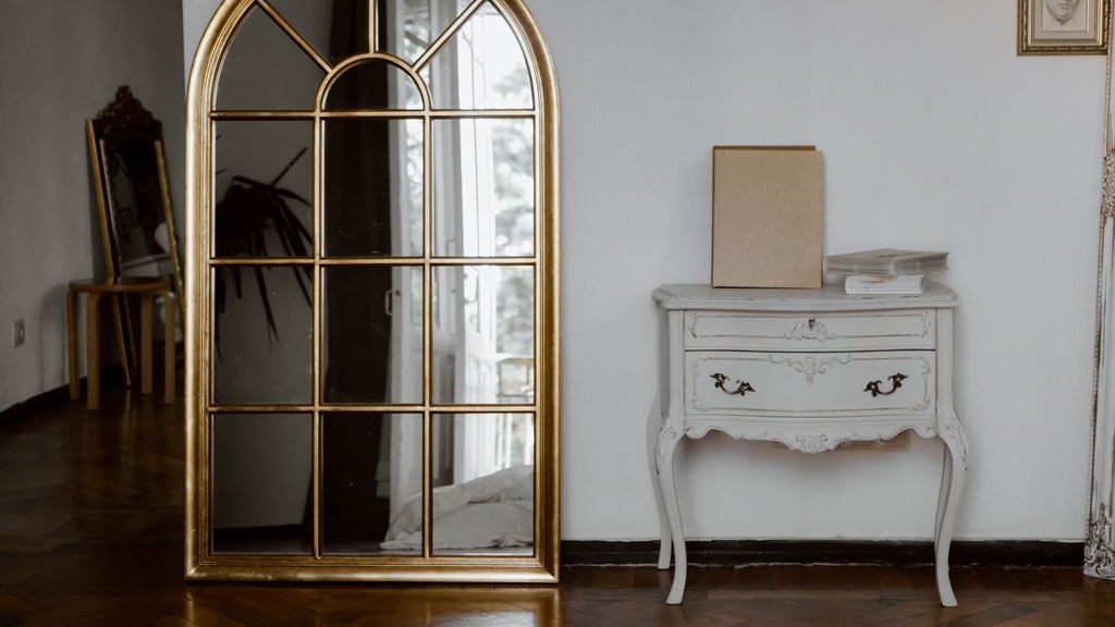 Vintage white console table with drawers beside a large arched gold mirror and framed wall art.