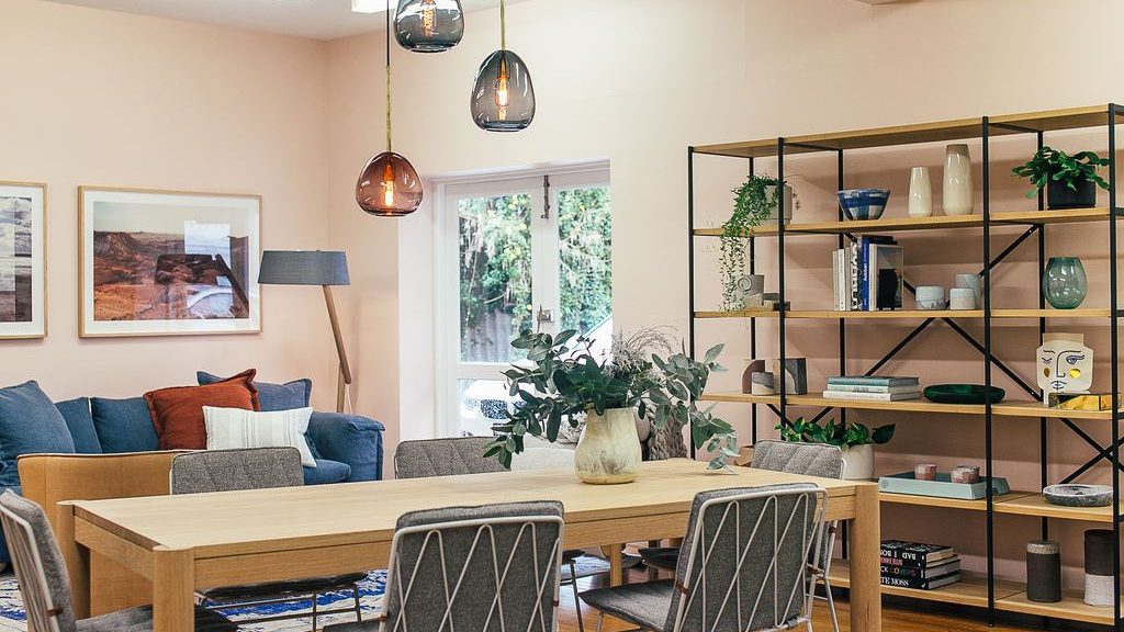 Open-plan Scandinavian-style dining and living room with a light wood table, gray chairs, indoor plants, open shelving, and colorful pendant lights.