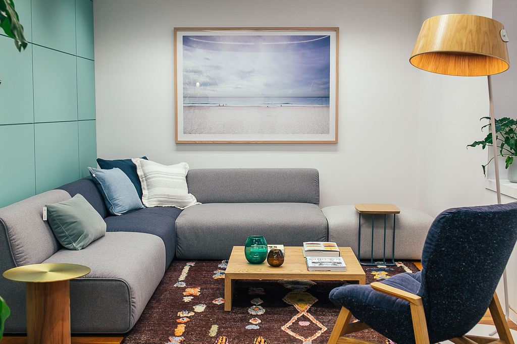 Modern coastal-inspired living room with gray sectional sofa, blue accent pillows, beach artwork, wood coffee table, and warm floor lamp.