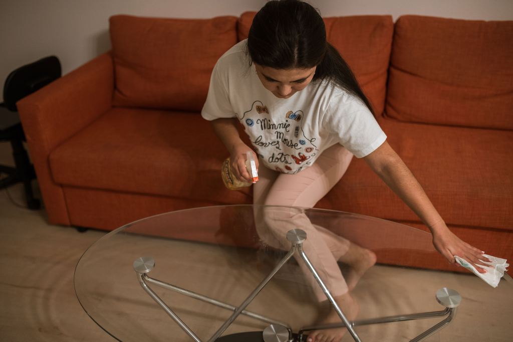 Woman wiping a round glass coffee table in front of an orange sectional sofa.