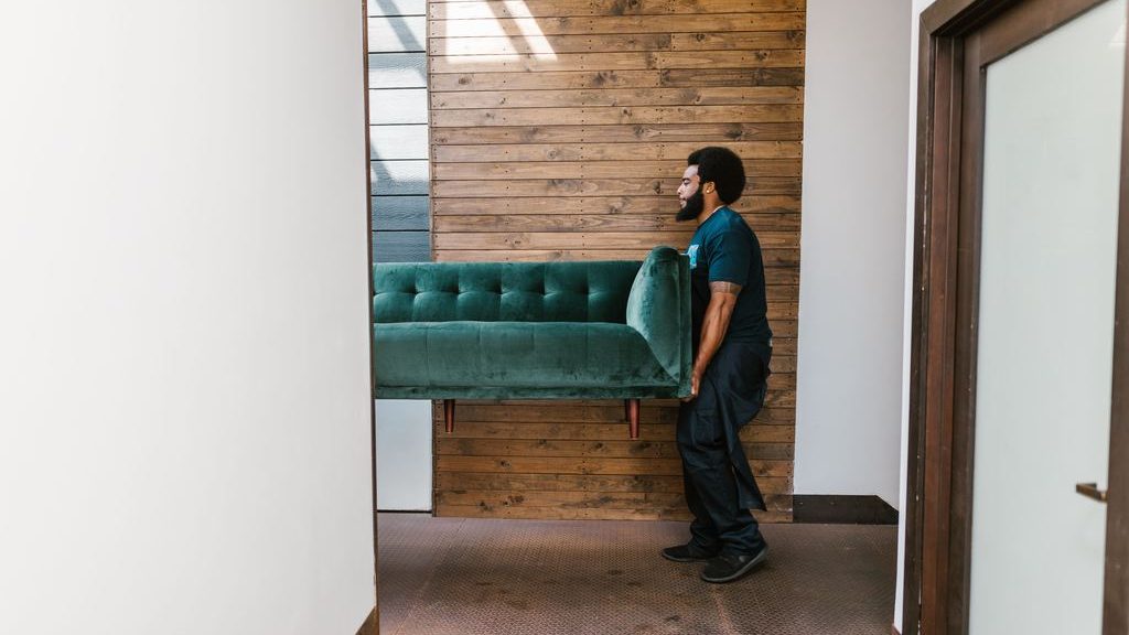 Man carrying a green upholstered sofa through a hallway during furniture delivery