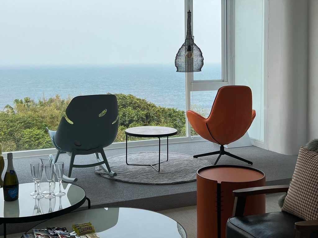 Contemporary coastal sitting area with ocean-view windows, colorful lounge chairs, round coffee tables, and soft neutral textures.