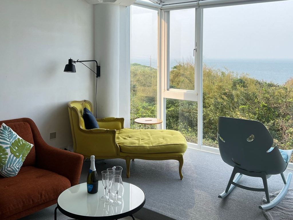 Cozy living room with a green chaise and rocking chair placed in front of floor-to-ceiling bay windows overlooking trees and the ocean.