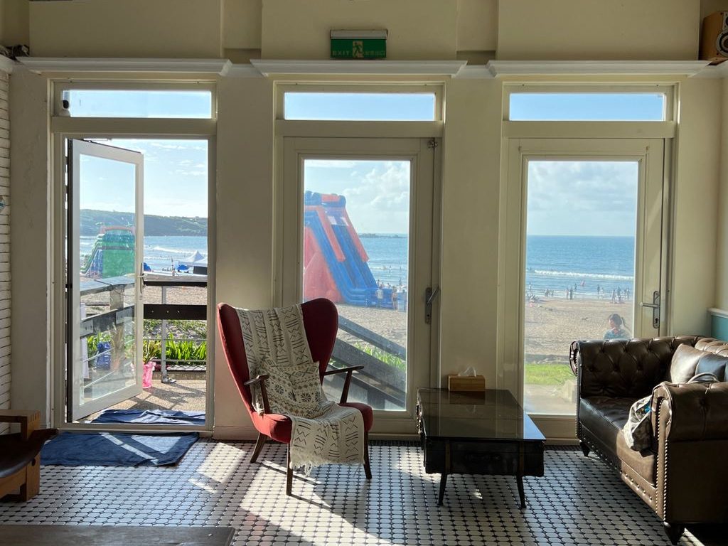 Sunlit coastal lounge with beach-view glass doors, leather sofa, red accent chair, patterned tile floor, and relaxed seaside seating.