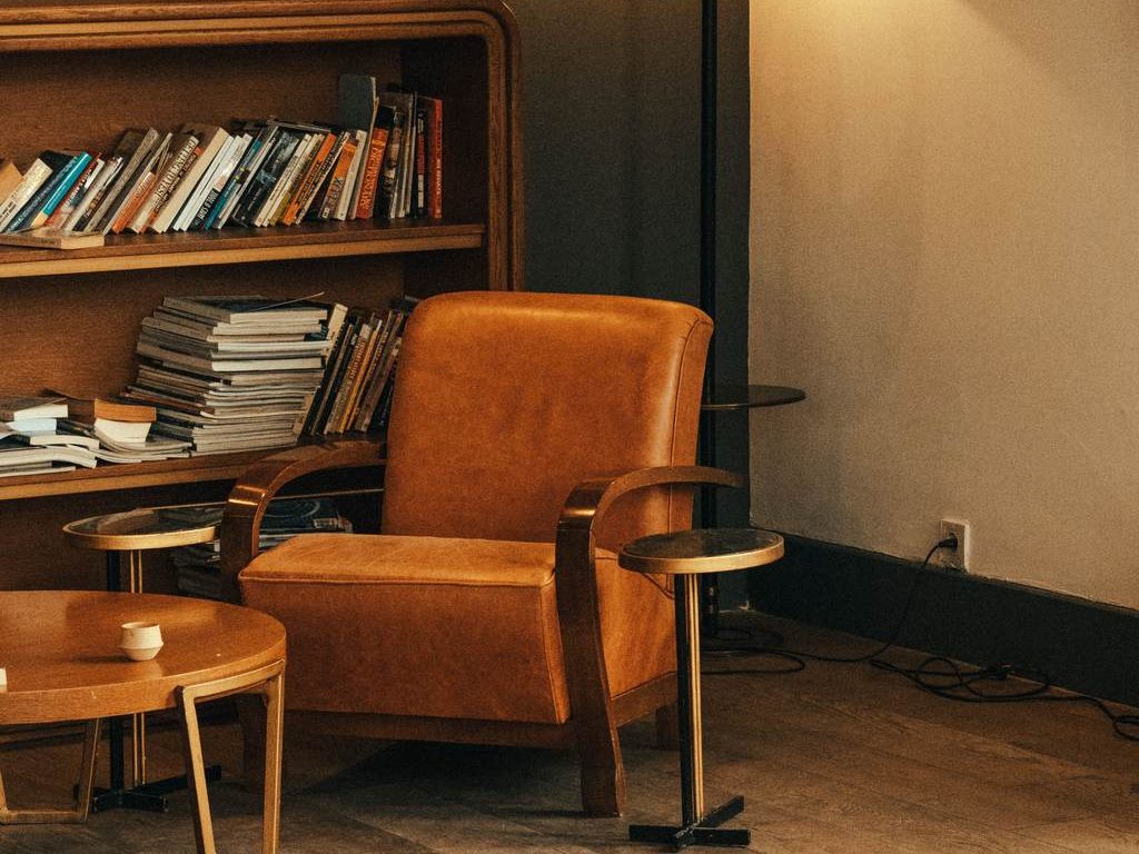 Leather reading chair beside a wood bookcase filled with books, floor lamp, small tables, and framed wall art in a warm living room.