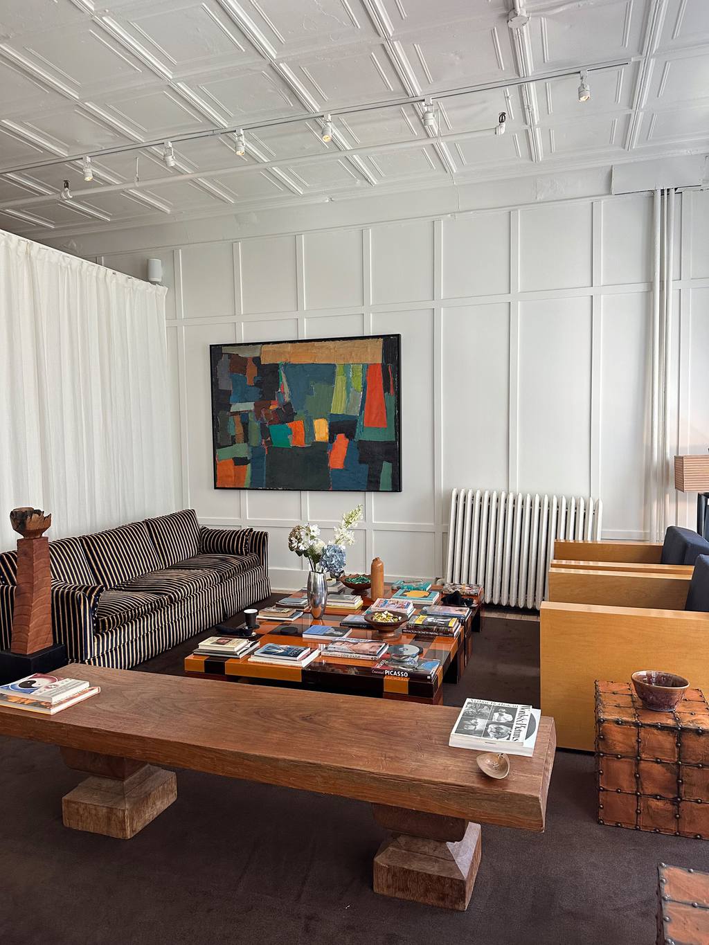 Eclectic living room with a striped sofa, oversized wooden bench, colorful abstract art, layered tables, and collected decor in a bright white-paneled space.