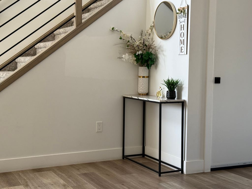 Slim black metal console table under a staircase, styled with a tall vase, small plant, round wall mirror, and entryway decor.