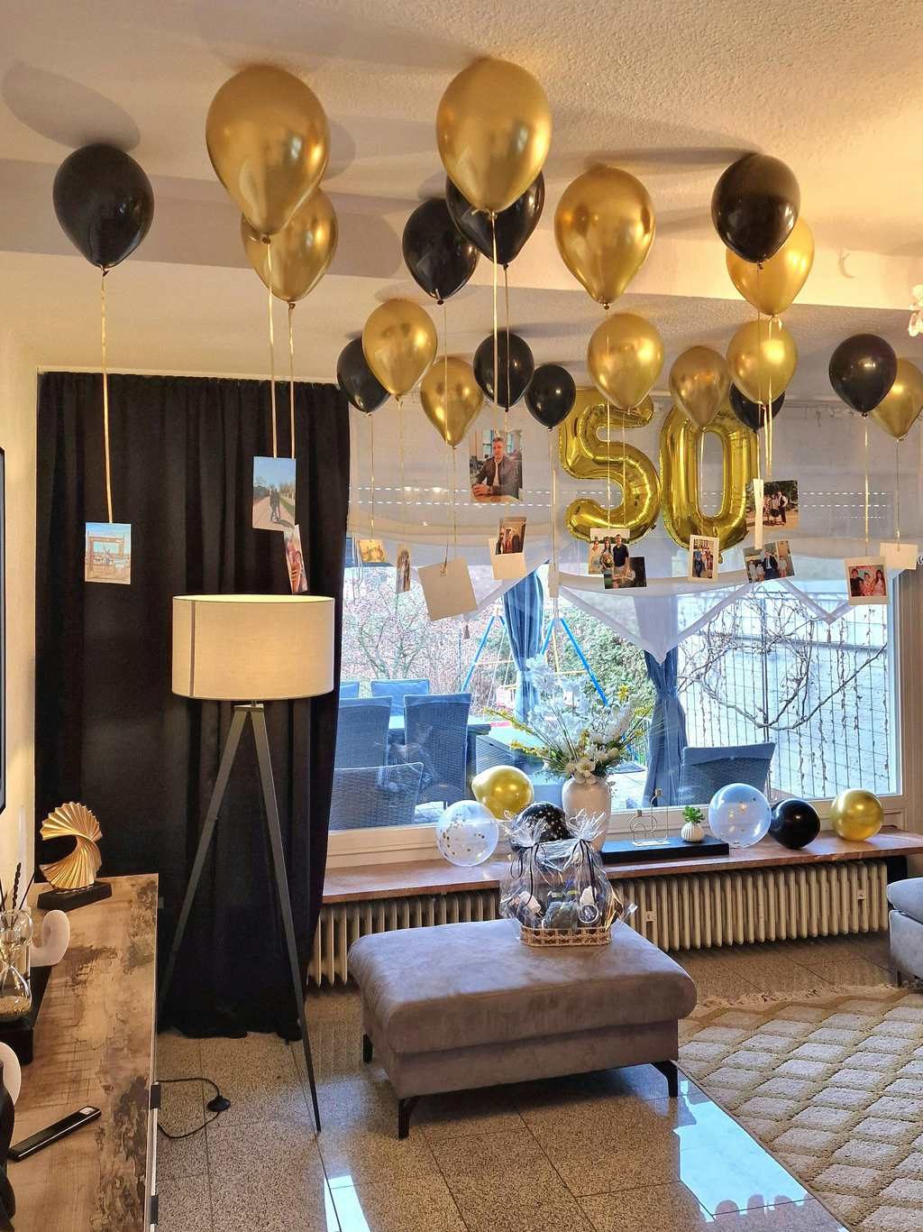 Living room decorated with black and gold balloons, hanging photo cards, large number balloons, and a gift basket for a festive indoor party setup.