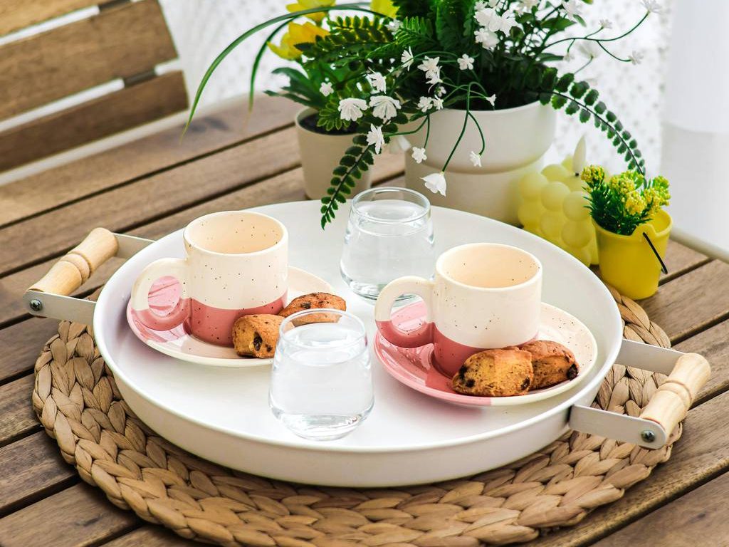 A natural organic tray setup with a woven base, white serving tray, greenery, and soft pastel drinkware on a wood table.