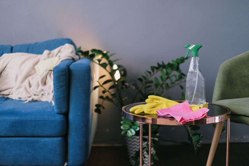 Cleaning gloves, cloth, and spray bottle on a side table beside a blue sofa in a living room.