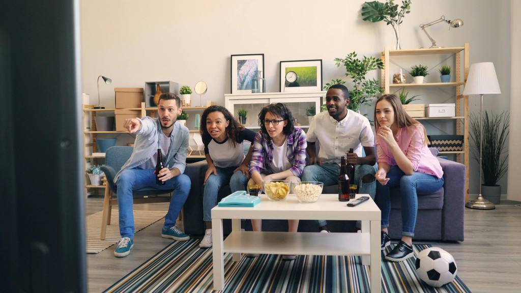 Friends watching a soccer match in a bright living room with snacks on the coffee table and a soccer ball near the sofa.
