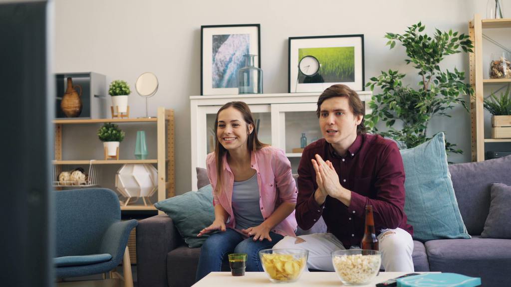 Couple watching a soccer match in a modern living room with snacks, drinks, sofa seating, plants, shelves, and cozy home decor.