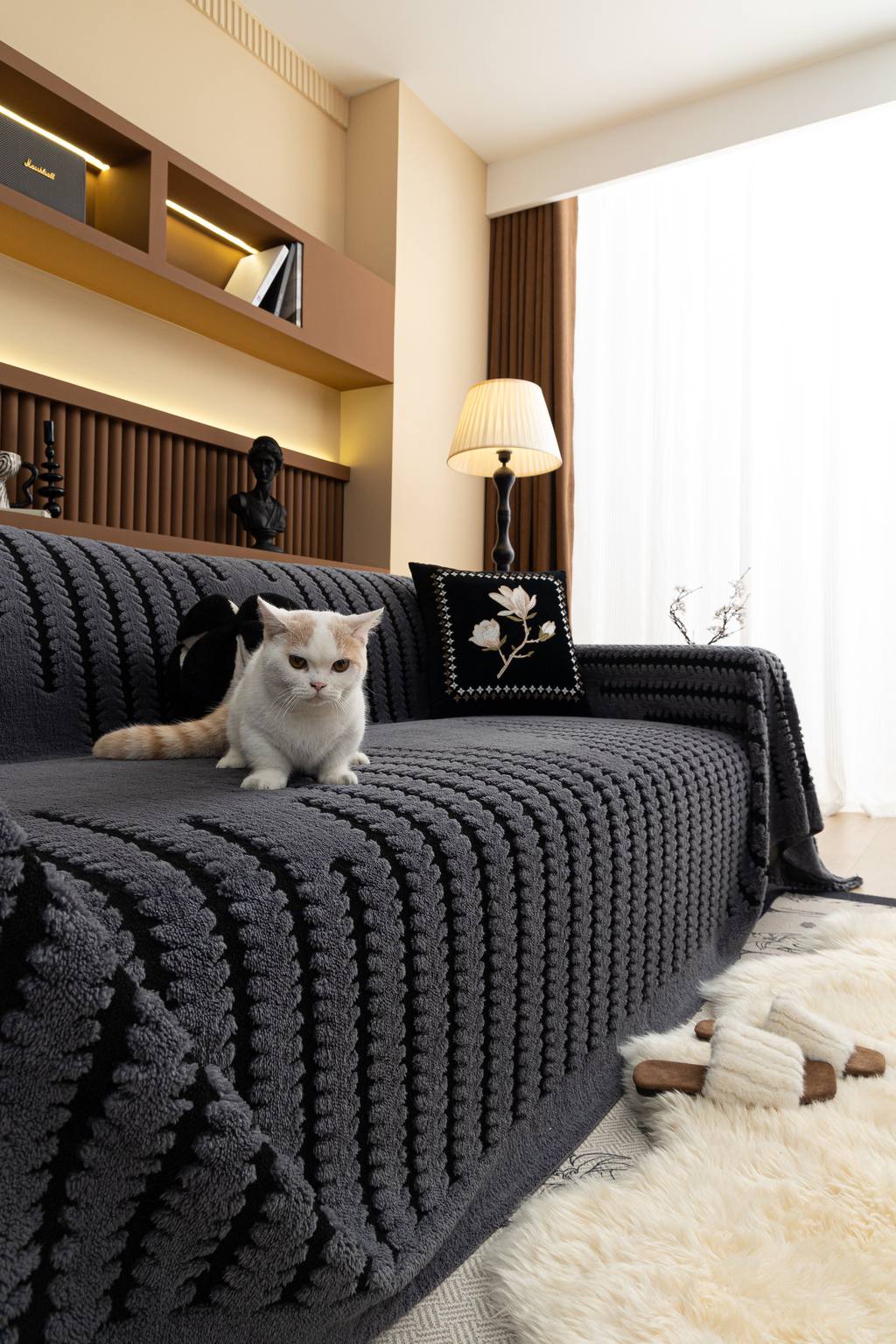 White-and-orange cat sitting on a dark textured sofa cover in a cozy living room with warm lighting, wood shelving, and a cream rug.