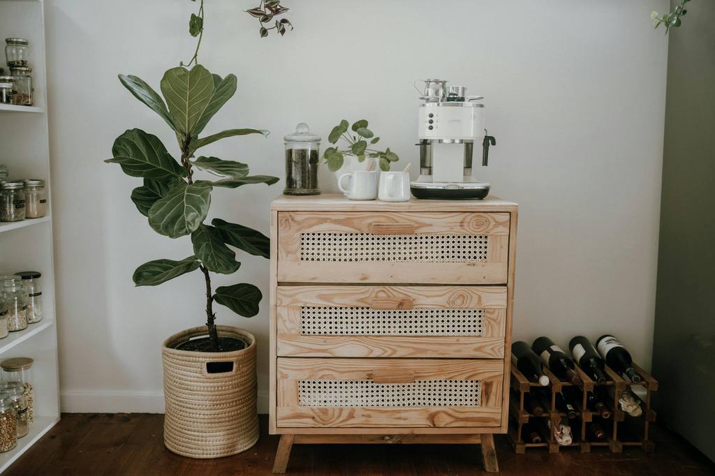 Light wood cabinet used as a home coffee bar with espresso machine, mugs, pantry jars, wine rack, and indoor plant.