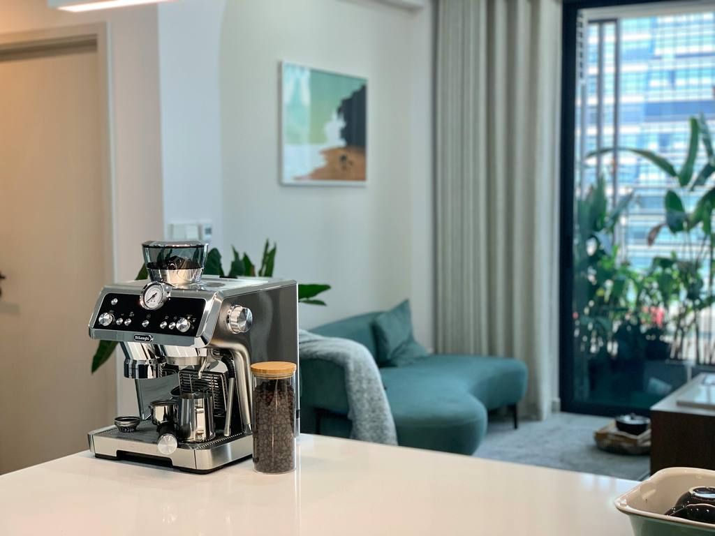 Modern espresso machine on a white kitchen counter with coffee beans, overlooking a bright living room with sofa, plants, and city window.