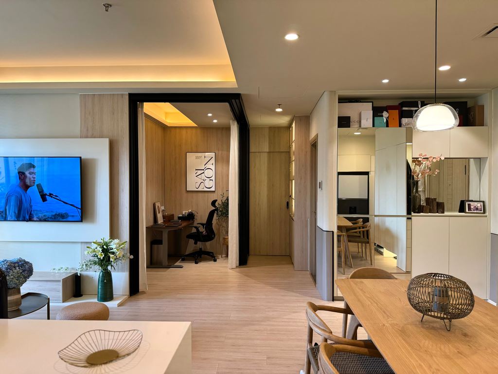 Open-plan living and dining space with a wall-mounted TV, compact home office nook, wood dining table, integrated storage, and warm recessed lighting.