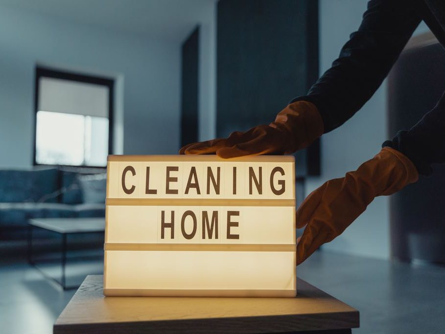 Gloved hands placing a “Cleaning Home” lightbox sign on a wooden table in a modern living room.