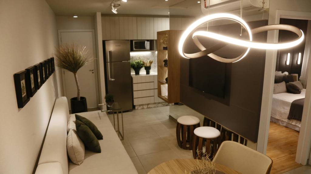 Small apartment living area with sofa, TV divider, compact kitchen, stools, and modern lighting arranged for efficient everyday use.