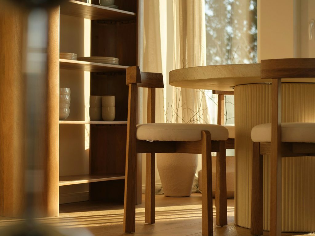 Sunlit dining room with a round table, wooden chairs, and open shelving styled with neutral ceramic dishes.