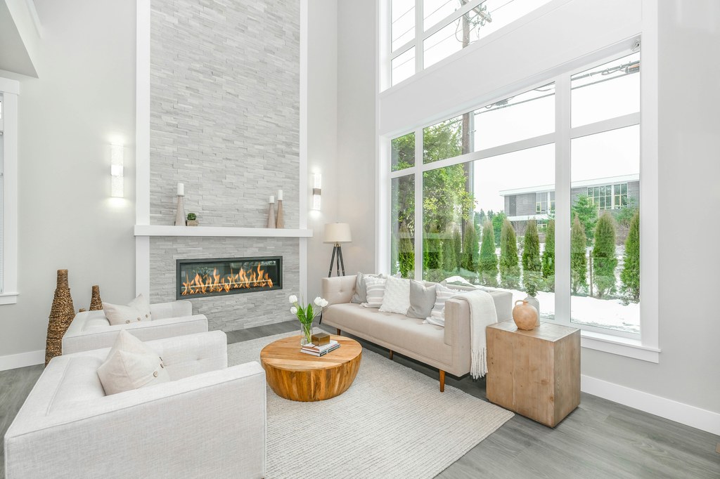 Bright modern living room with a beige sofa placed in front of floor-to-ceiling windows, white armchairs, fireplace, and round wood coffee table.