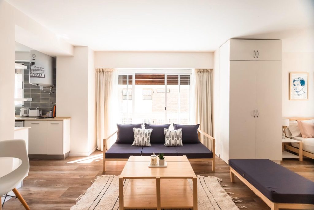 a large living room with sofa, wood coffee table and an ottoman next to a kitchen and a bedroom in a minimalist and compact apartment