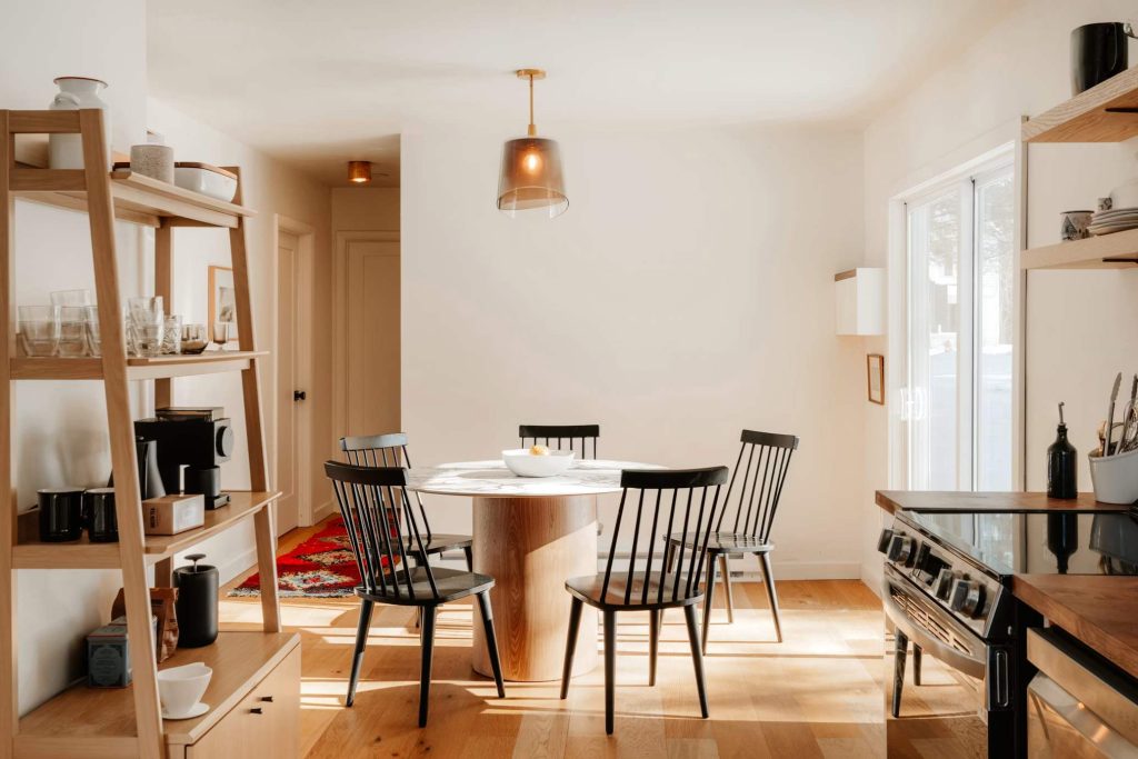 a round dining table with marble top, wood leg and five chairs near a wood shelf with glasses and decorations in a minimalist dining room