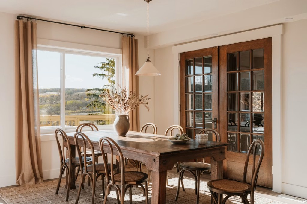 Warm traditional dining room with rustic wood table, curved-back chairs, neutral curtains, pendant light, and wooden French doors.