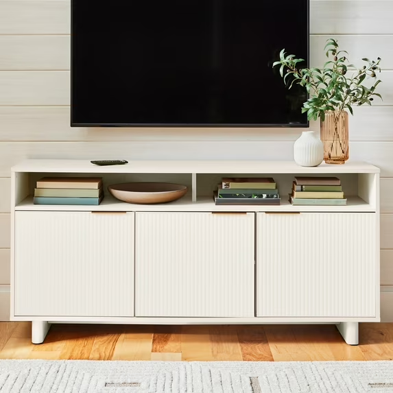 Better Homes & Gardens Lillian white Fluted TV Stand with storage for books and CDs and a vase and a plant on top against a white wall with a mounted tv in a living room