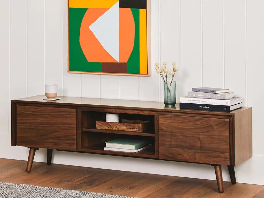 a wood rectangular media console with books, a candle and a vase on top against a white wall with a mounted painting in a modern living room