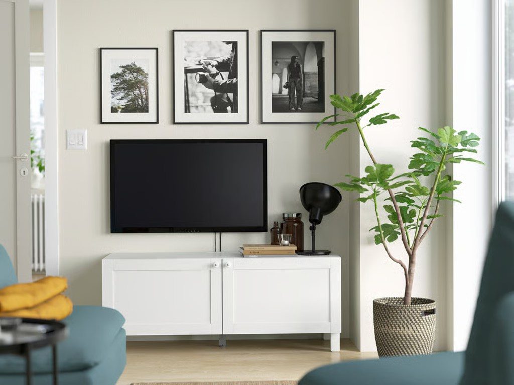 a white media console with a lamp and decorations on top below a wall-mounted tv near a plant and sofa in a modern and minimalist living room