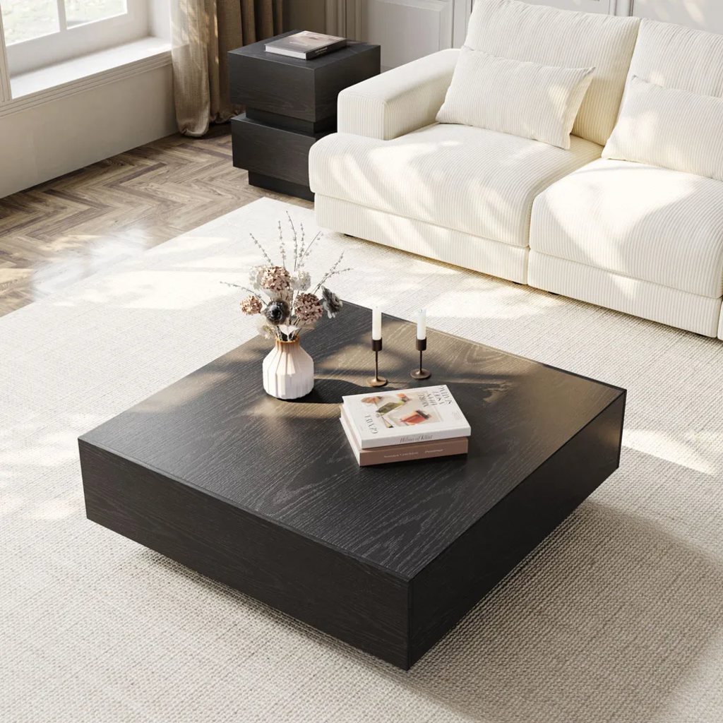 a rectangular black coffee table with storage and some books, two candles and a vase on top near a sofa and a side table in a minimalist living room