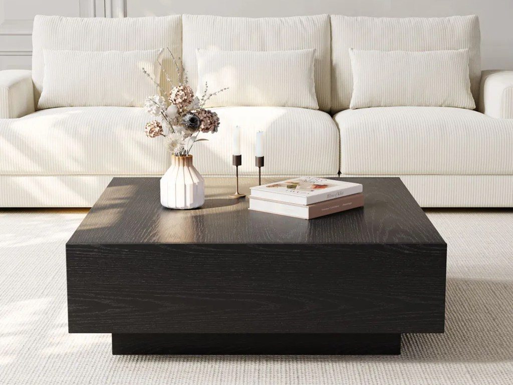 a black rectangular coffee table with two books, two candles and a vase on top near a beige tufted sofa in a bright living room