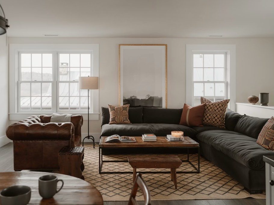 a rectangular wood coffee table with some books and decorations on top near a brown vintage leather sofa and a black chaise sofa in the center of the living room