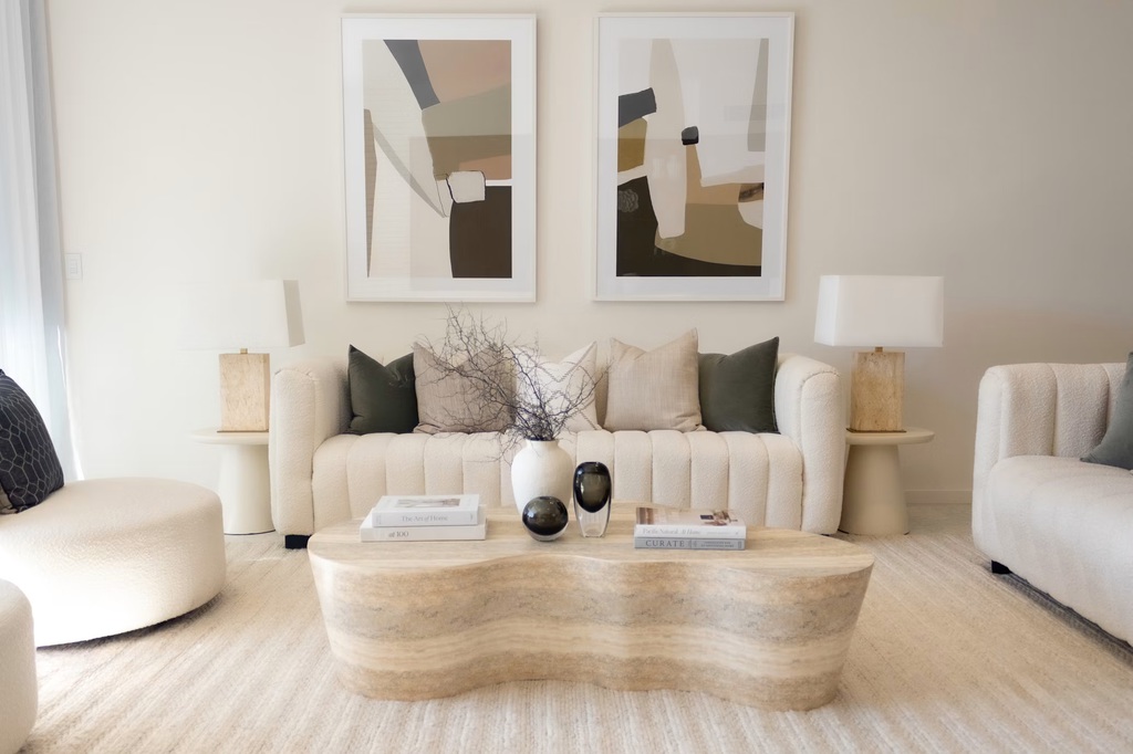 Cream transitional living room with curved sofa, abstract wall art, stone coffee table, neutral rug, and soft modern lounge chairs.