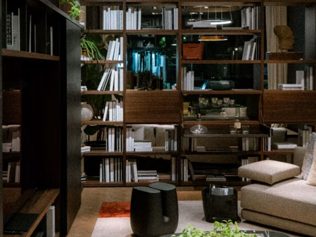 Cozy modern living room with dark wood bookshelves, neatly placed books, lounge seating, black coffee table, plants, and warm layered lighting.