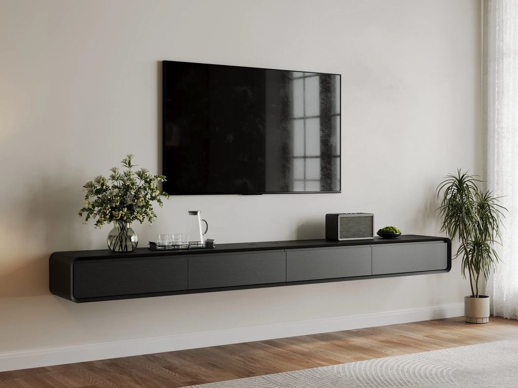 Wall-mounted TV above a long black floating media console, styled with plants and small decor in a bright modern living room with wood flooring and a light patterned rug.