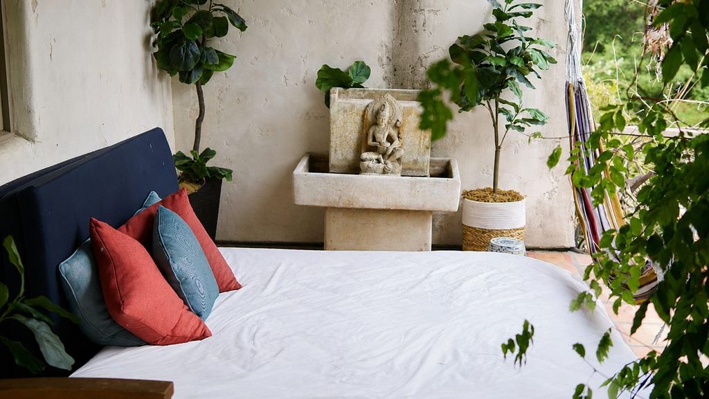 Sunlit boho-style bedroom with a white bed, colorful pillows, lush indoor plants, and a small stone fountain against a textured plaster wall.