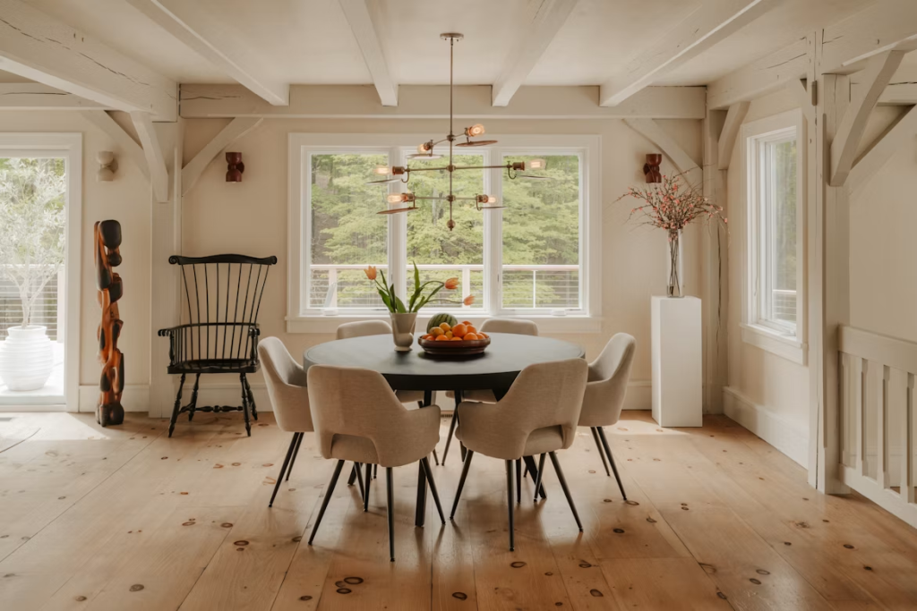 Elegant black round dining table surrounded by beige upholstered chairs in a rustic, high-ceiling open home.